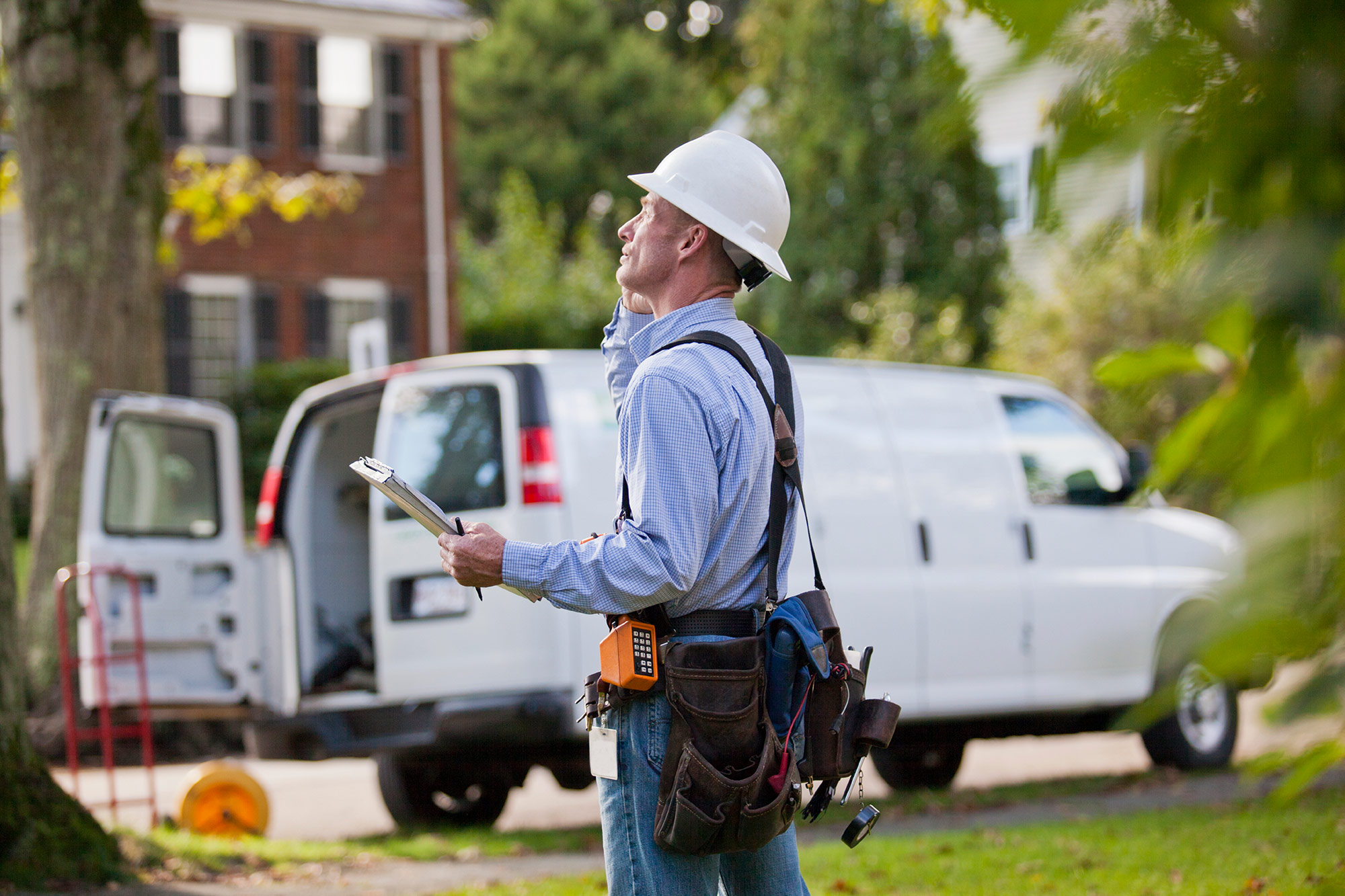 Man wearing a hard hat and tool belt with work vehicle in the background.