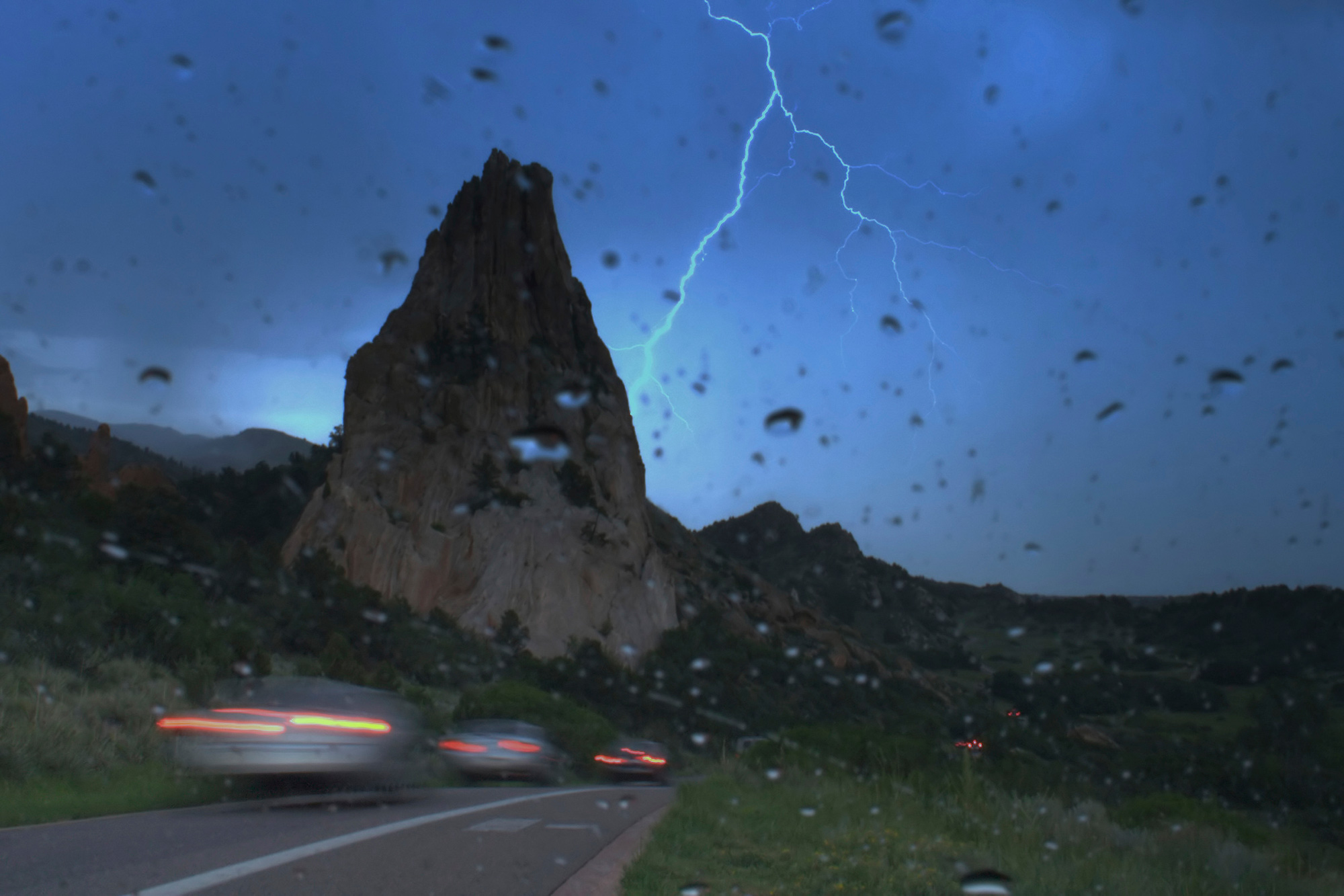 Blury cars driving on mountain road visable from a behind a vehicle's rainy windshield.