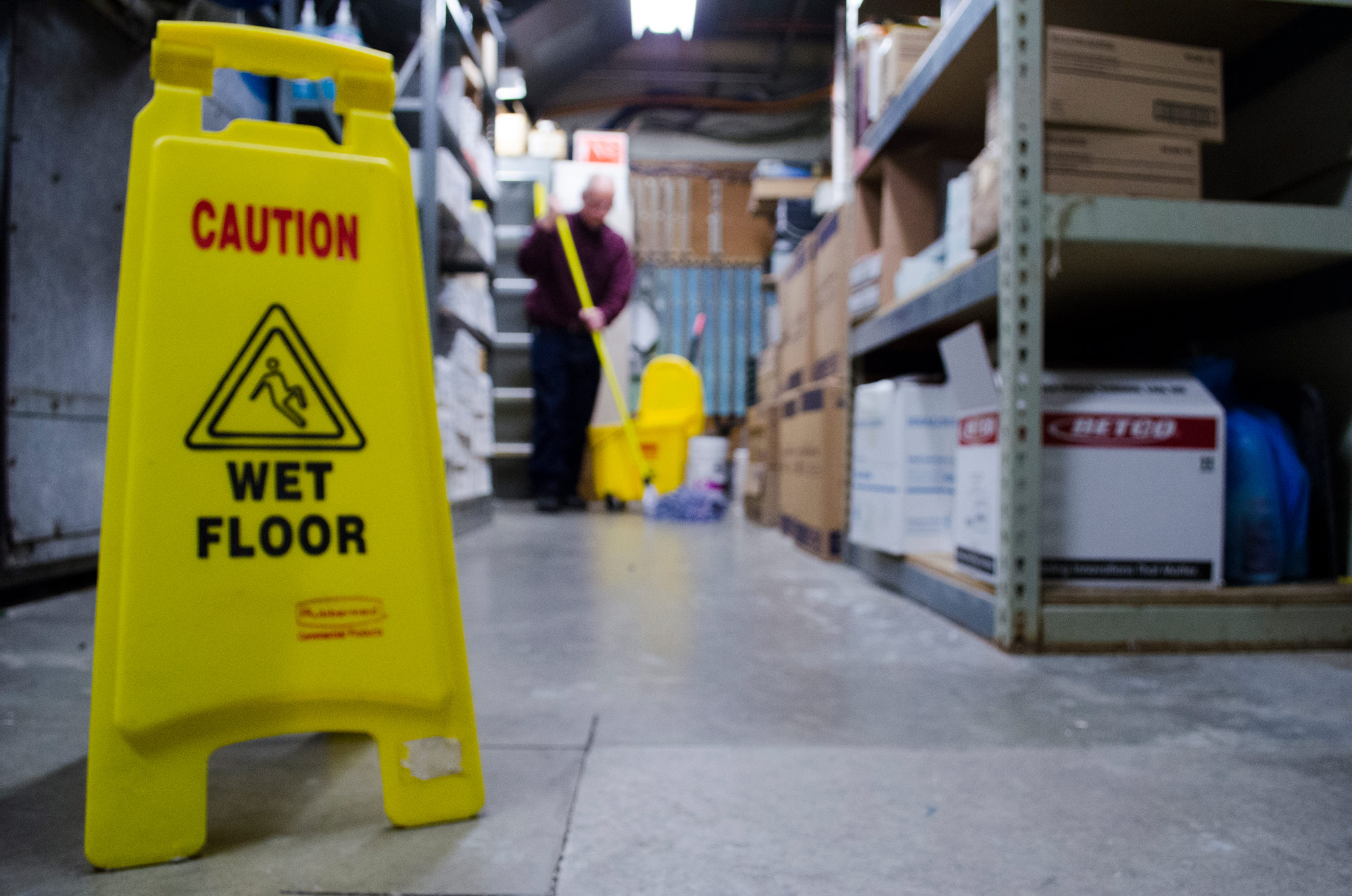 Yellow Caution - Wet Floor Sign in the foreground with a man mopping a warehouse floor in the background.