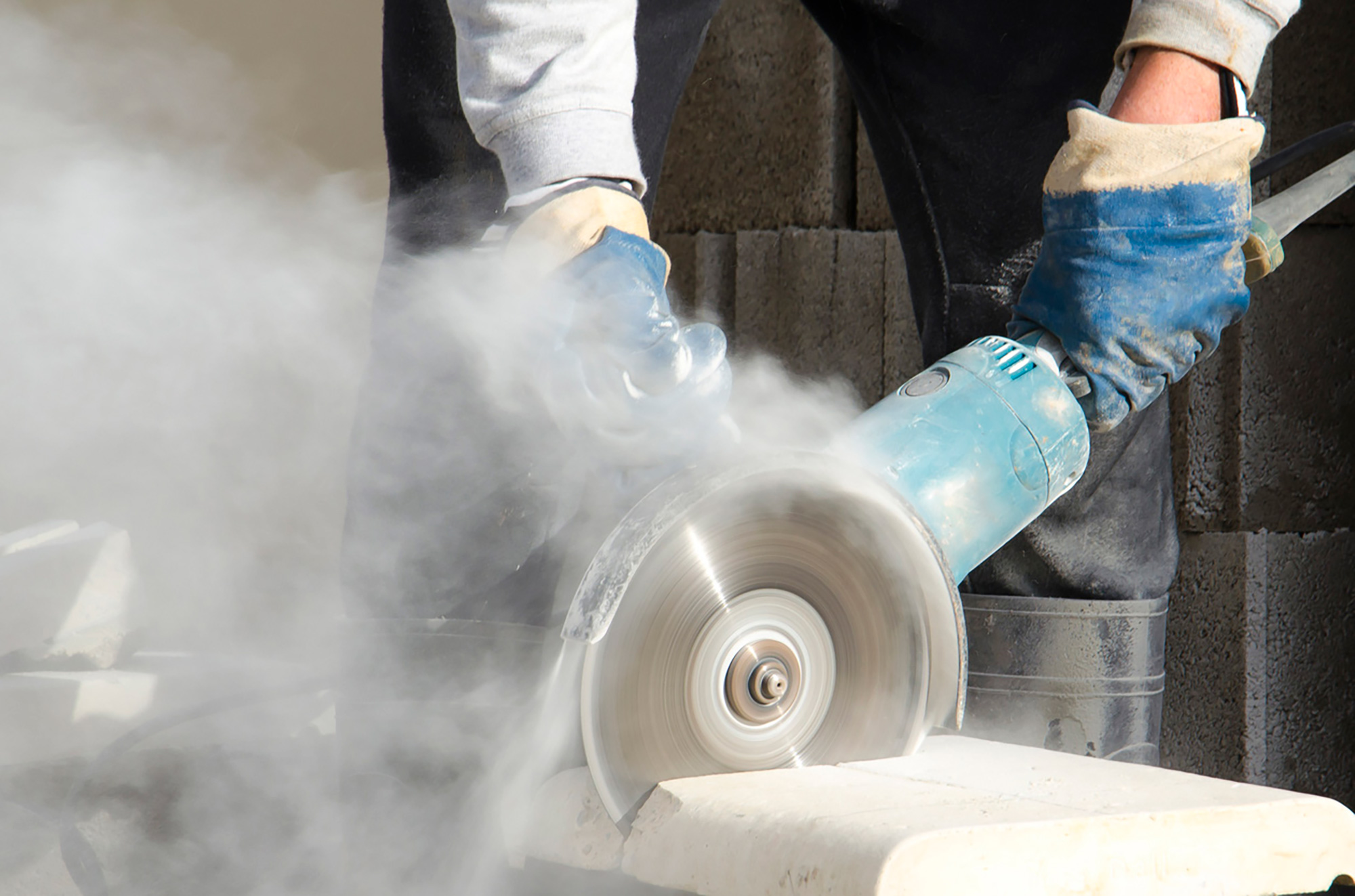Person using a circular saw to cut stone-link material with a cloud of white dust.