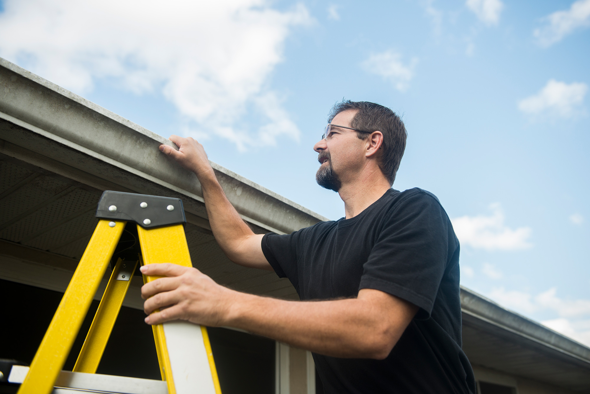 Man on ladder inspects the roof of his home.
