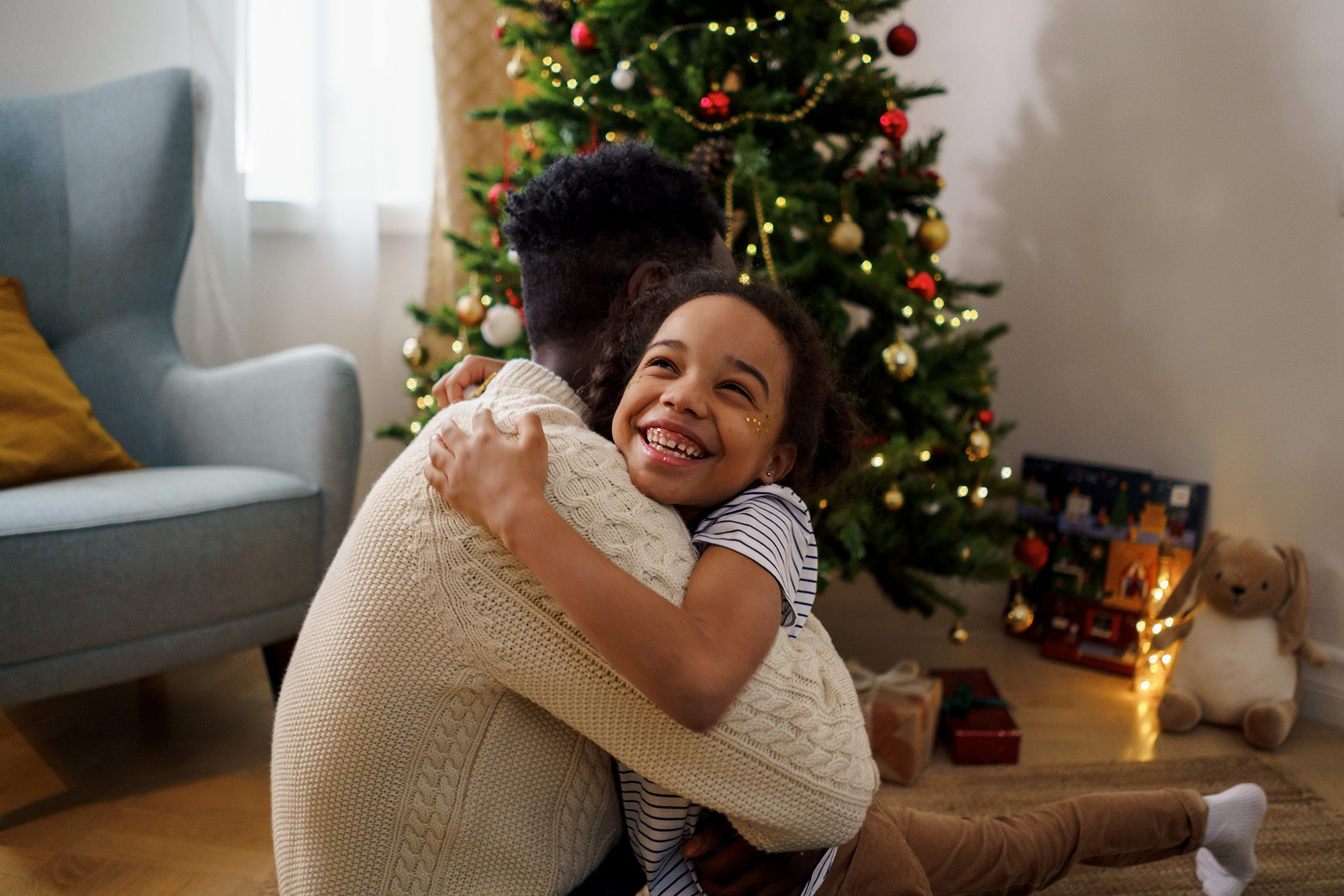 Father and daughter hug in front of decorated holiday tree.