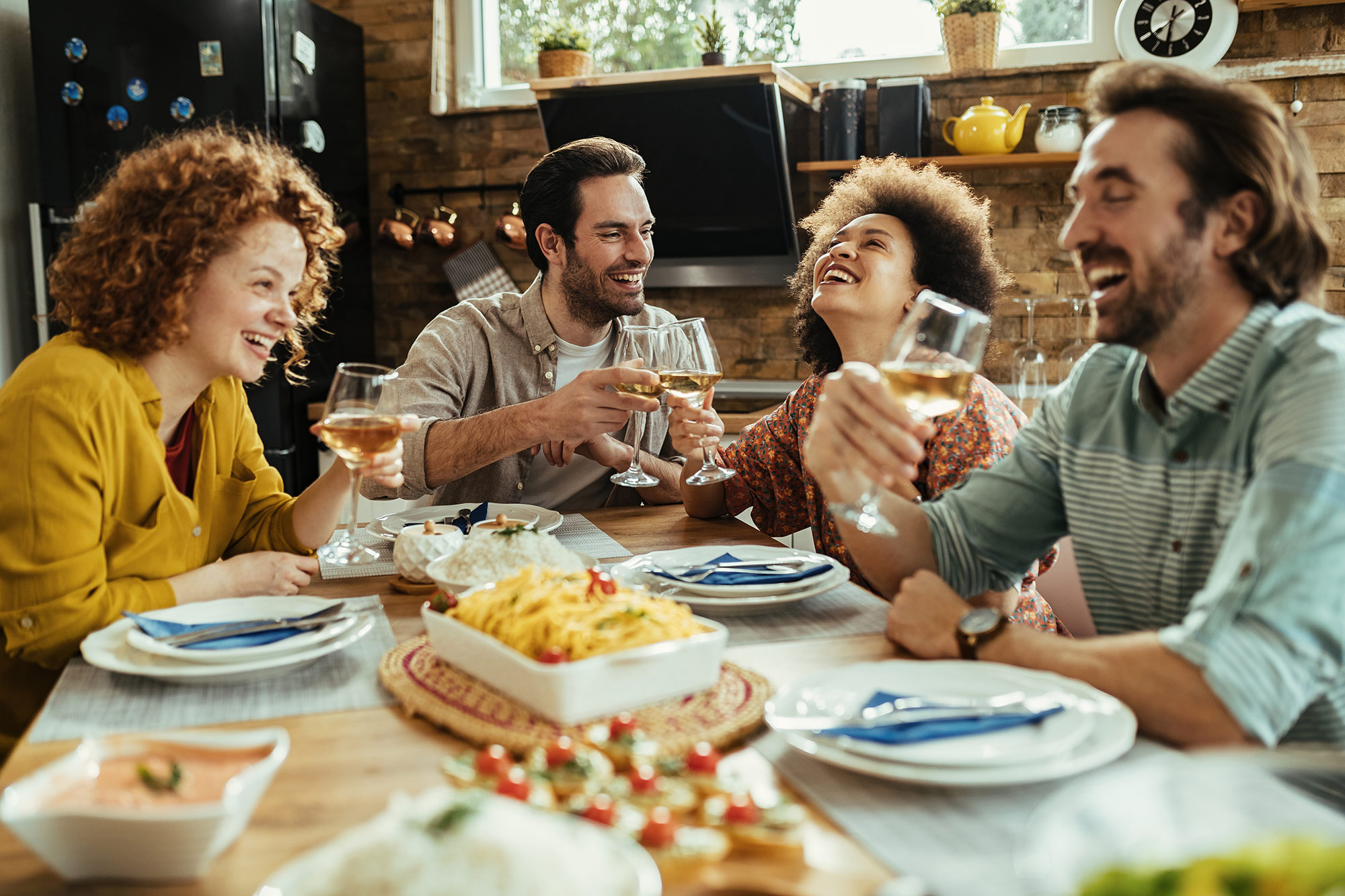 Group of four friends eating, drinking, and laughing together in a home.