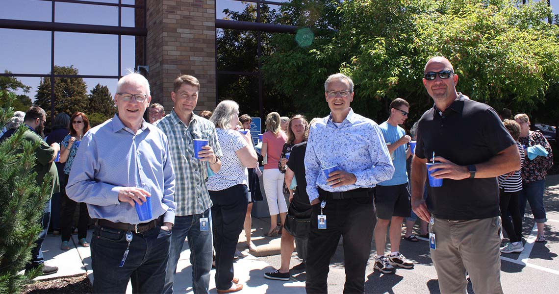 Four Western National employees holding blue Solo cups posing together outdoors on a sunny spring day with other employees mingling in the background.