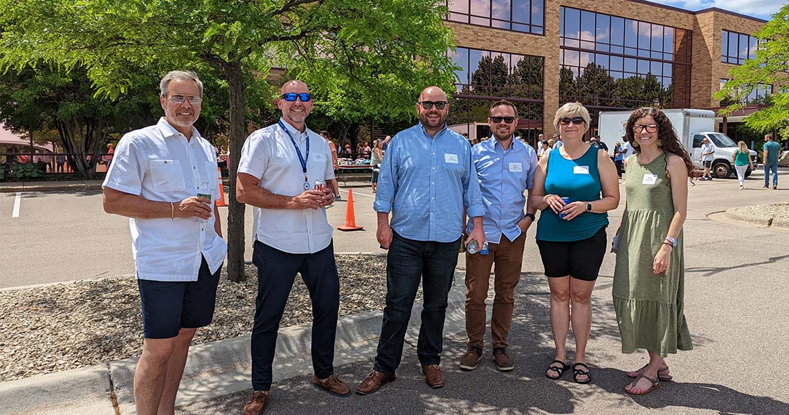 Six Western National employees wearing sunglasses posing together outdoors on a sunny spring day with the exterior of Western National's Edina office building in the background.