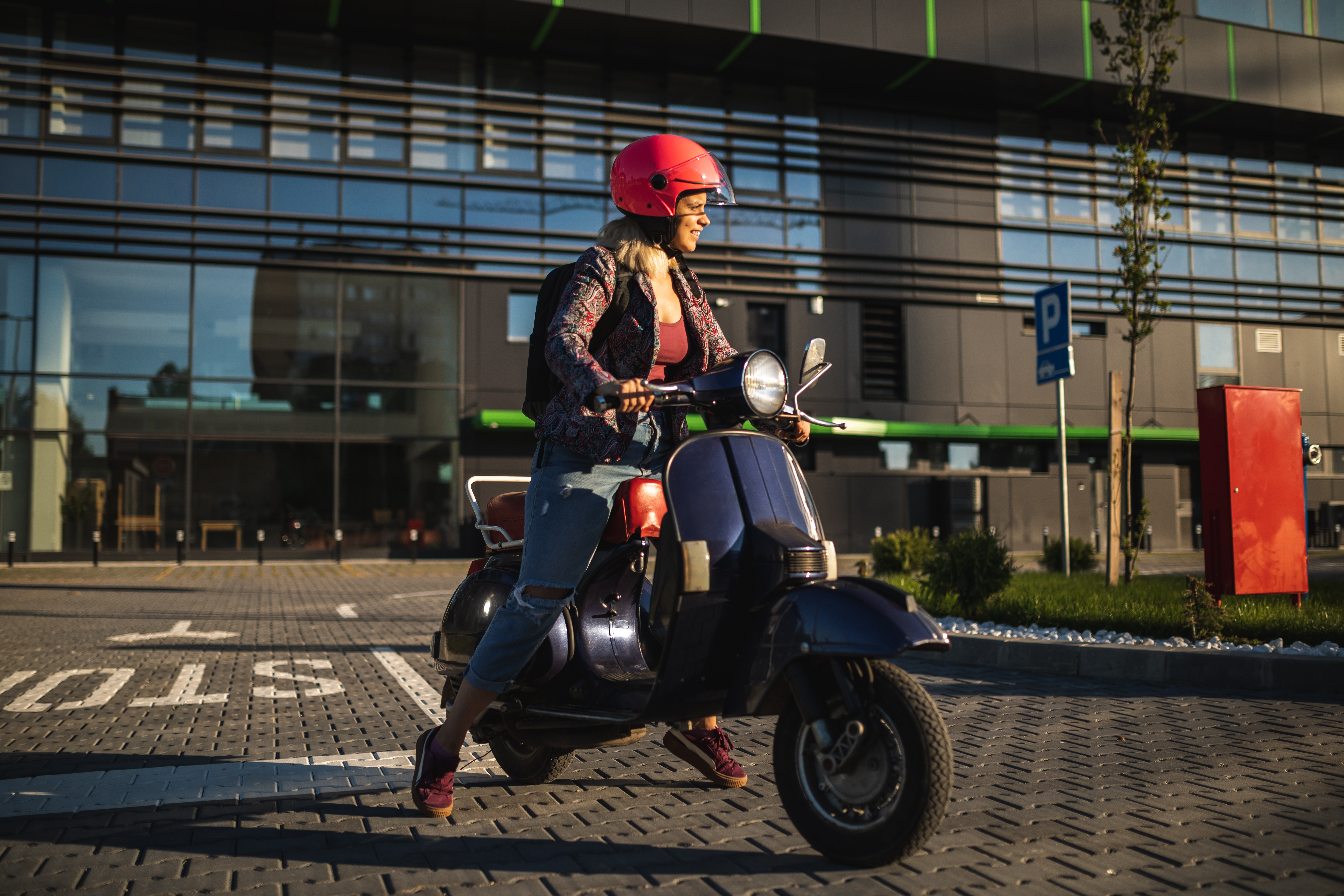 Woman in a red helmet sitting on a scooter in an urban parking lot.