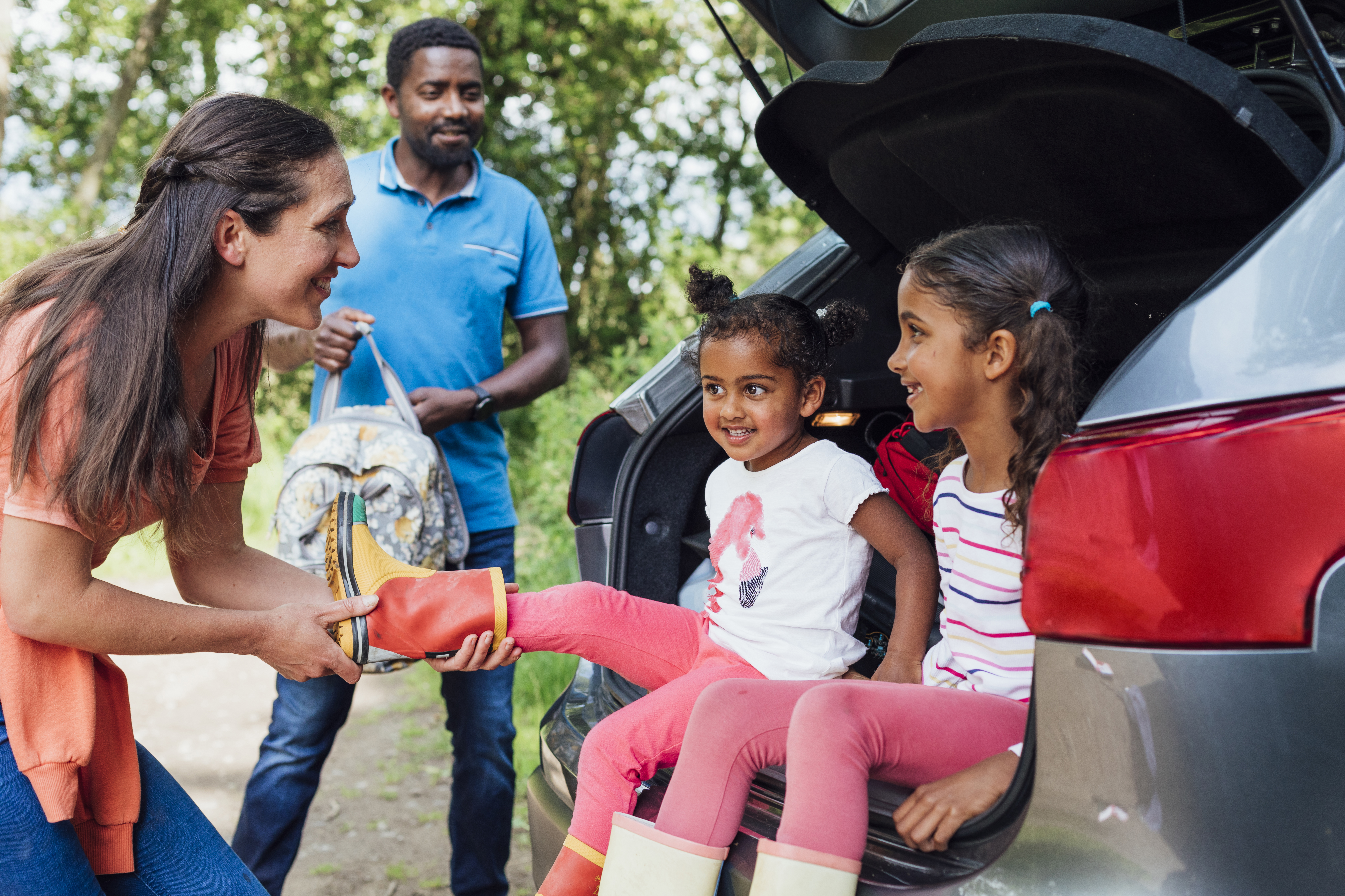 A woman helps a young child put on a red rain boot as two children sit in the open trunk of a car and another adult stands nearby holding a bag.